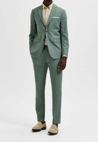 Man i en skräddarsydd ljusgrön kostym med en beige skjorta och ljusbeige loafers, stående med en hand i fickan.
