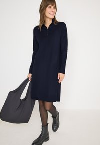 Abito in maglia blu navy con colletto con zip, orlo a costine e maniche lunghe. Abbinato a stivali neri con lacci e a una morbida borsa a tracolla nera.