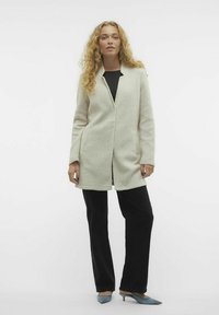 Cappotto di lana grigio chiaro con apertura frontale, tasche laterali e vestibilità comoda, abbinato a pantaloni neri e tacchi a punta blu.