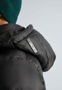 Schwarze gepolsterte Jacke mit hohem Kragen und einem Logo-Patch mit der Aufschrift "JACKIT" auf der Schulter. Der Stoff wirkt glatt und glänzend.