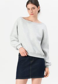 Giovane donna in felpa grigia chiara a spalle scoperte e gonna mini in denim scuro, in piedi contro uno sfondo bianco.