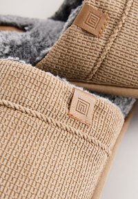 Beige Stoffhausschuhe mit strukturierter Außenseite, grauem, flauschigem Futter und einem kleinen Lederetikett mit geometrischem Logo.