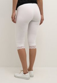 Witte capri leggings van rekbare stof, met kantafwerking aan de zoom en een elastische tailleband. Gepaard met witte sneakers.