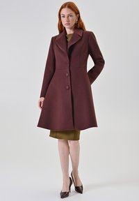Cappotto in misto lana bordeaux con corpino aderente, ampio colletto e tre bottoni frontali; indossato sopra un vestito aderente verde oliva e abbinato a tacchi alti.