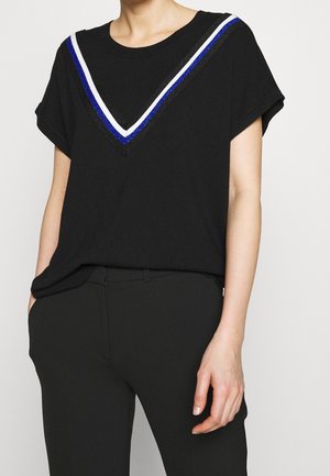 Femme portant un haut noir à manches courtes avec des rayures en forme de V bleues, blanches et noires au niveau de l'encolure, associé à un pantalon noir.