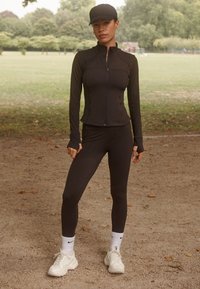 Giacca atletica nera con collo alto e zip, abbinata a leggings neri. Scarpe da ginnastica bianche e calzini con logo Nike. Sfondo di un parco.