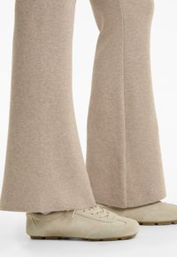 Beige suède veterschoenen gecombineerd met lichtbruine, wijde gebreide broeken die over de schoenen vallen, op een witte achtergrond.