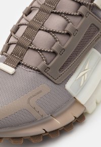 Närbild av taupe- och beige Reebok-sneakers med texturerat tyg, förstärkta snöröglor, mönstrade snören och slitstarkt yttersula.