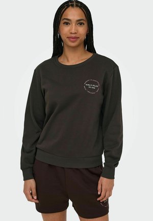 Ung kvinde med fletninger bærer mørk sweatshirt og matchende shorts med små runde hvide tekstlogoer på venstre bryst og ben.