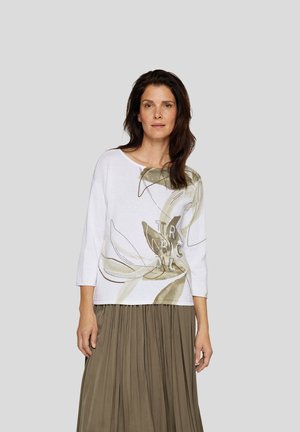 White, loose-fitting top with three-quarter sleeves, featuring a green and beige floral design and the text "TROPICAL." Worn with a pleated skirt.