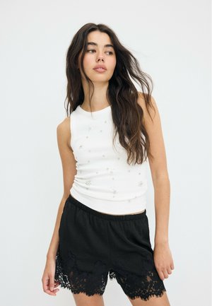 Ung kvinde med langt mørkt hår iført en hvid ærmeløs top med små pynt og sorte shorts med sort blonder mod en hvid baggrund.