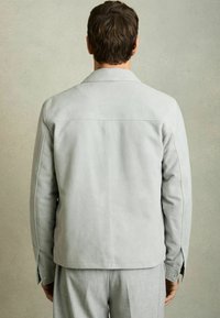 Jachetă din suede gri cu o textură netedă, cu manșete cu nasturi și un guler clasic, văzută din spate, purtată peste pantaloni gri deschis.