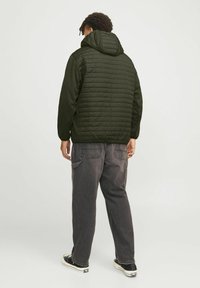 Veste matelassée vert foncé avec une capuche, arborant un design matelassé. Portée avec un pantalon gris ample et des baskets noires.