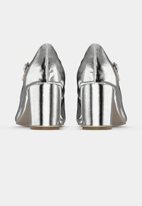 Zapatos de tacón alto plateados con un acabado brillante, puntera redondeada y tacón ancho; presentan un detalle de hebilla en el costado.