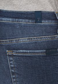 Vaqueros de denim azul oscuro con un tejido texturizado, con costuras en contraste, un bolsillo trasero y una etiqueta de tela azul marcada con '7'.