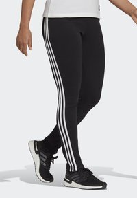 Svart leggings med tre vertikale hvite striper på sidene, laget av elastisk materiale, sammen med svarte treningssko.