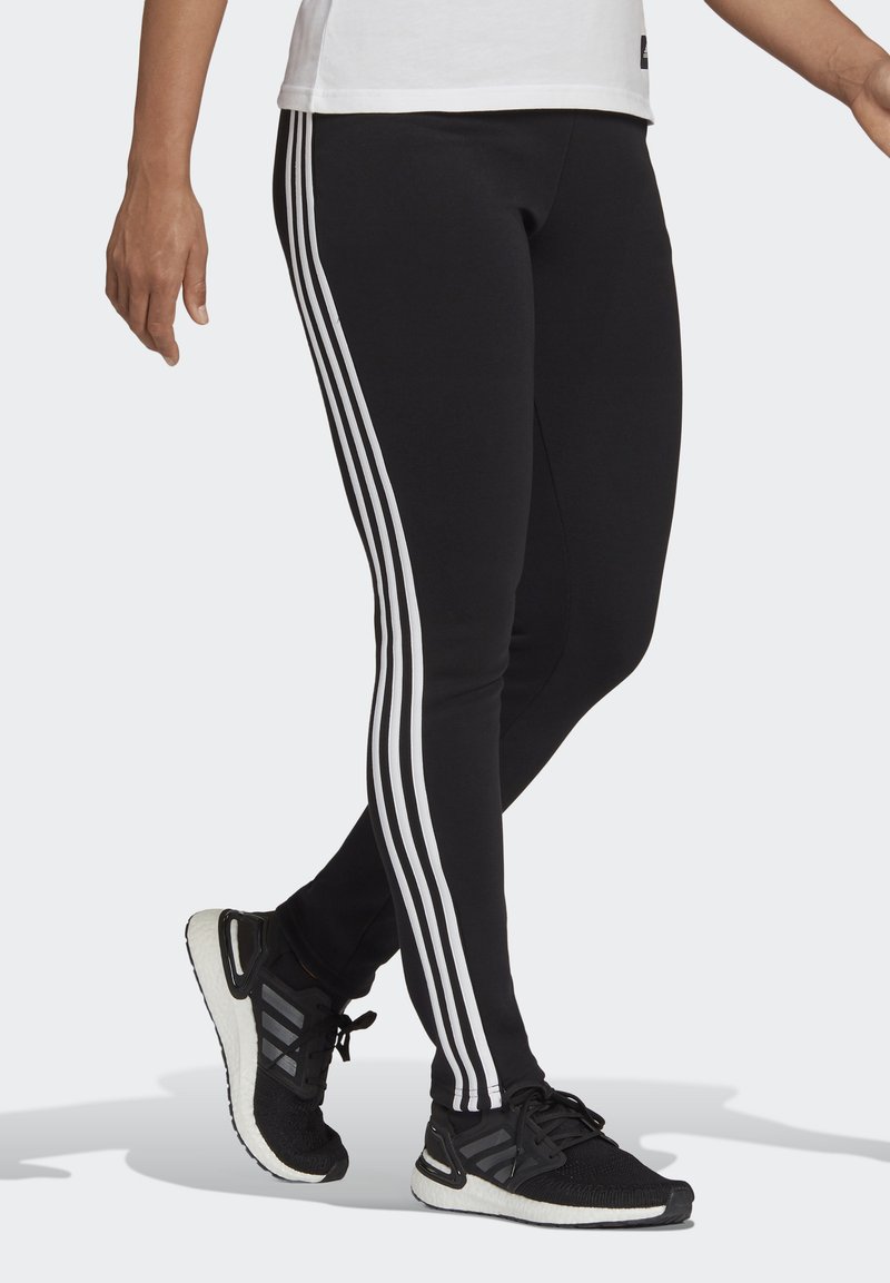 Svart leggings med tre vertikale hvite striper på sidene, laget av elastisk materiale, sammen med svarte treningssko.