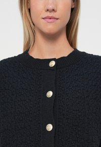 Marine teksturert strikket cardigan med ribbestrikket hals og tre gullfargede knapper foran.