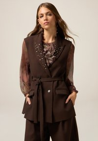 Bruine op maat gemaakte mouwloze blazer met versieringen, een ceintuur in de taille en twee zakken, gedragen over een doorschijnende blouse met bloemenpatroon.