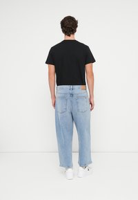 Μαύρο βαμβακερό t-shirt, ανοιχτό μπλε χαλαρές τζιν παντελόνια με καφέ δερμάτινη πατίνα στη μέση, λευκά αθλητικά παπούτσια, βλέπεται από πίσω.