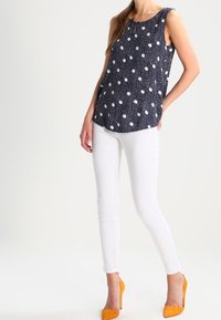 Marineblauw mouwloos topje met witte stippen, gecombineerd met witte strakke broeken en oranje hoge hakken. Gladde stof en een losse pasvorm.