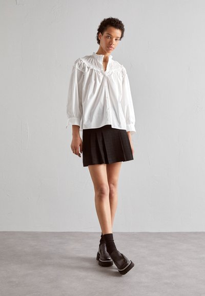 Hvid, langærmet bluse med smock-detaljer ved kraven, parret med en plisseret sort miniskirt og kraftige sorte sko.