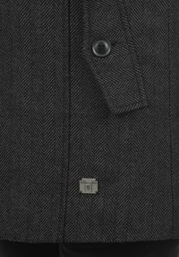 Černý kabát se vzorem rybí kosti, zapínáním na knoflíky, texturovanou látkou a malou kovovou značkou, zdůrazňující střih s vertikálními pruhy.