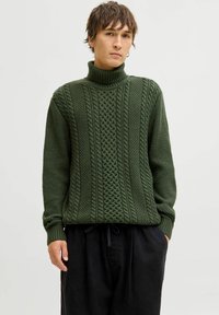 Un pull vert foncé avec un col roulé haut, un motif en relief de torsades et des poignets côtelés. Porté avec un pantalon noir ample.