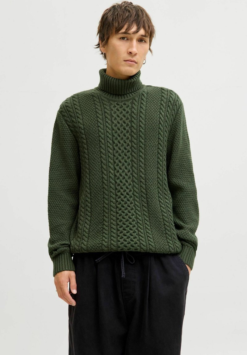 Un pull vert foncé avec un col roulé haut, un motif en relief de torsades et des poignets côtelés. Porté avec un pantalon noir ample.