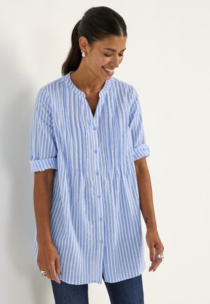 Frau lächelt, trägt ein blau-weiß gestreiftes Hemd mit Knöpfen und hochgekrempelten Ärmeln, steht vor einem schlichten hellen Hintergrund.