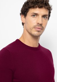 Bordeauxrode trui gemaakt van zachte stof, met een ronde hals en een gladde textuur. Close-up van het bovenlichaam en gezicht van een model.