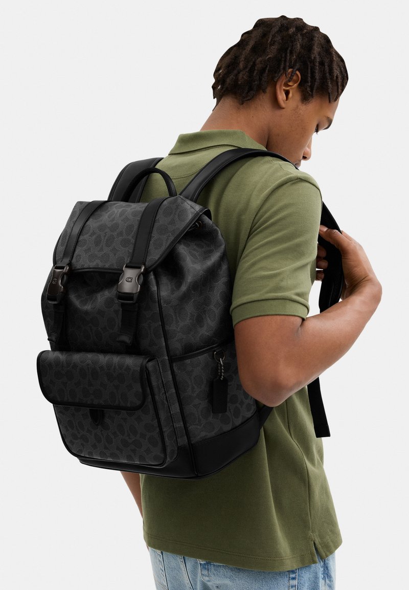 Hombre con polo verde oliva y vaqueros azul claro ajustando las correas de una mochila negra con estampado, bolsillo delantero y cierres con hebillas.
