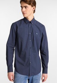 Camicia blu navy a maniche lunghe con un piccolo motivo geometrico, colletto a bottoni e logo ricamato sul petto. Tessuto in cotone, texture liscia.
