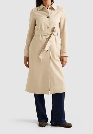 Kvinde iført en beige trenchcoat med knapper og bundet bælte, mørkeblå vidde jeans og brune sko mod en ensfarvet baggrund.