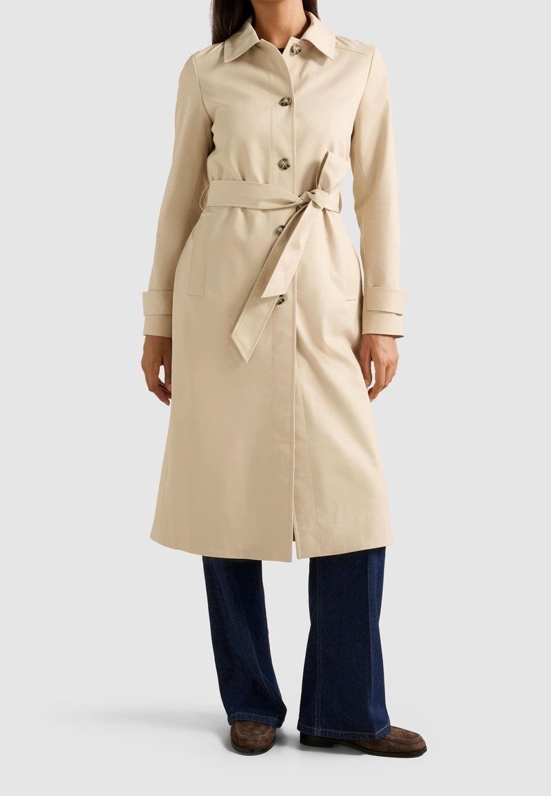 Femme portant un trench-coat beige avec des boutons et une ceinture nouée, un jean large bleu foncé et des chaussures marron sur un fond uni.