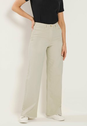 Donna che indossa pantaloni beige a gamba larga, top nero a maniche corte, sneakers bianche, con una mano sui fianchi e l'altra rilassata lungo il corpo.