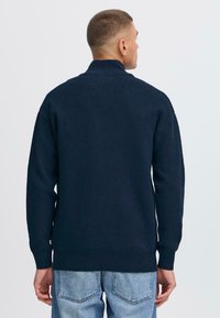 Pullover lavorato a maglia blu navy con colletto alto, polsini e orlo a coste. Tessuto liscio; dettagli delle cuciture visibili sulle spalle e sui lati.