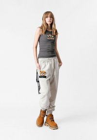 Camiseta sin mangas gris con detalle bordado, pantalones de chándal beige con un parche de oso y botas de tobillo marrones con suela rugosa.