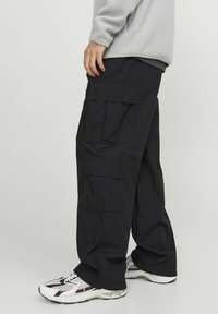 Pantaloni cargo neri realizzati in tessuto resistente. Presentano ampie tasche laterali, una vestibilità ampia e un design a gamba dritta. Indossati con scarpe da ginnastica bianche e nere.