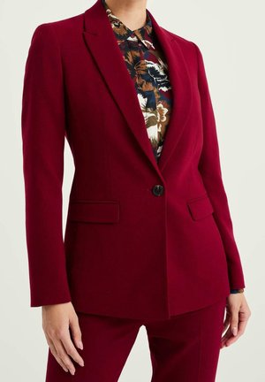 Femme portant un blazer rouge foncé cintré et un pantalon assorti, associé à une chemise fleurie à boutons, se tenant devant un fond uni.