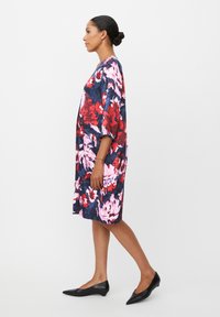 Bloemenjurk in marineblauw, rood en roze met oversized mouwen. Glad materiaal, kniehoogte, met een gedurfde bloemenprint. Draagend met zwarte flats.