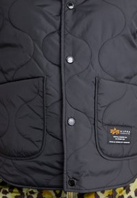 Veste matelassée foncée avec deux poches avant, boutons-pression de marque et un petit patch rectangulaire Alpha Industries sur la poche inférieure droite.