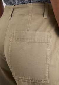 Khaki aukštos juosmens kelnės, turinčios lygią tekstūrą, su užpakaline kišene ir dvigubai siūtu siūlu, kad būtų užtikrintas didesnis patvarumas.