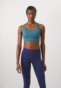 Sostén deportivo acanalado en color verde azulado con tirantes estrechos, combinado con leggings morados de cintura alta que presentan una sutil textura y variación de color.