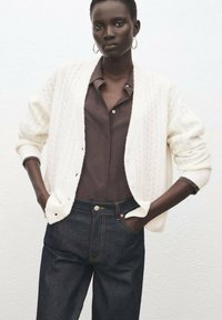 Cárdigan blanco de punto con textura de trenzas, combinado con una camisa de botones marrón oscuro y vaqueros de mezclilla oscuros. Pendientes sencillos de aro metálico.