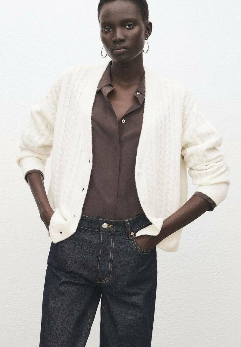 Cárdigan blanco de punto con textura de trenzas, combinado con una camisa de botones marrón oscuro y vaqueros de mezclilla oscuros. Pendientes sencillos de aro metálico.