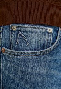 Närbild av en blå denimjeans bakficka med metallnitar och en brun ribbad tröja instoppad.