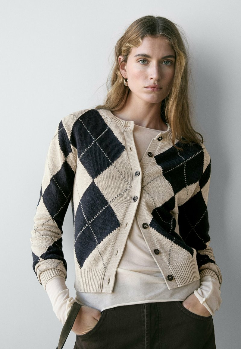Jonge vrouw met golvend haar draagt een beige en zwarte argyle cardigan over een licht shirt, staand met haar handen in de zakken van donkere broek.