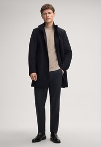 Donker wollen overjas met hoge kraag, gecombineerd met een lichtbeige trui en donkere gestructureerde broek. Glad leren schoenen maken de outfit af.