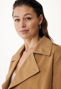 Cappotto trench beige con colletto a rever, realizzato in un tessuto liscio. Dettagli notevoli includono una leggera cucitura a contrasto e orecchini a cerchio dorati.